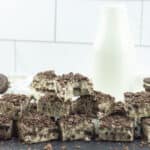 A stack of cookies and cream dessert bars made with a rich cookies and cream fudge recipe is topped with crushed cookies. A glass bottle of milk and whole sandwich cookies rest in the background against a white tiled wall.