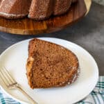 A slice of rich chocolate Bundt cake sits on a white plate with a fork, resting on a gray surface beside a green and white striped cloth, while the remaining chocolate Bundt cake awaits on a wooden stand in the background.