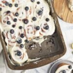 A baking pan filled with frosted cinnamon rolls topped with fresh blueberries—perfect for a Mother's Day tea party. One roll is missing, revealing the soft interior. Nearby are a plate with a roll, loose blueberries, and a lemon wedge.