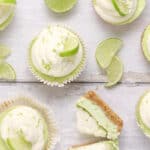Lime cupcakes with white frosting and lime wedges are arranged on a light wooden surface, reminiscent of mini key lime cheesecakes. Some cupcakes are whole, while one is cut to show the inside. Lime slices and zest are scattered around.