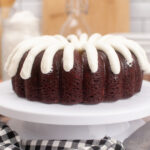 A chocolate bundt cake with thick white cream cheese frosting draped over the top, displayed on a white cake stand—a festive addition inspired by classic Christmas bundt cake recipes. Eggs, a checkered towel, and a glass of milk sit nearby on the kitchen counter.