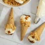 Three waffle ice cream cones filled with whipped cream and topped with crumbles and bits of dried fruit rest on parchment paper, reminiscent of peach cobbler cheesecake cones. A piping bag and a bowl of crumbles are nearby on a white surface.