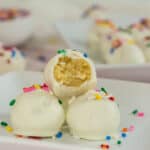 Three white chocolate cake balls with colorful sprinkles on a plate, one with a bite taken out showing a soft, moist center—the perfect graduation dessert idea. More cake balls and sprinkles are blurred in the background.