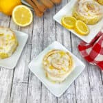 Three lemon-glazed cinnamon rolls on white plates, garnished with lemon zest, are arranged on a wooden surface with lemon halves, a red and white checkered cloth, and wooden utensils—perfect for Easter dessert ideas.