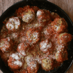 A black skillet filled with zucchini meatballs in tomato sauce, sprinkled with grated cheese, sits on a wooden surface—a delicious twist on the classic zucchini meatballs recipe.