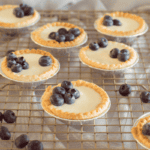 Several mini cheesecakes topped with fresh blueberries sit on a cooling rack, reminiscent of a Lemon Blueberry Tart. The cheesecakes are in foil cups and the scene has a warm, inviting atmosphere, with extra blueberries scattered around.