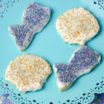 Four cookies decorated with white icing and colorful sprinkles, shaped like fish and circles, are arranged on a turquoise lace-edged plate. Perfect as summer snacks for kids, the plate sits on a colorful, scale-patterned background.