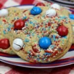 A plate with festive cookies topped with red, white, and blue candies and sprinkles, perfect for 4th of July Recipes for Kids, sits on a red and white checkered tablecloth.