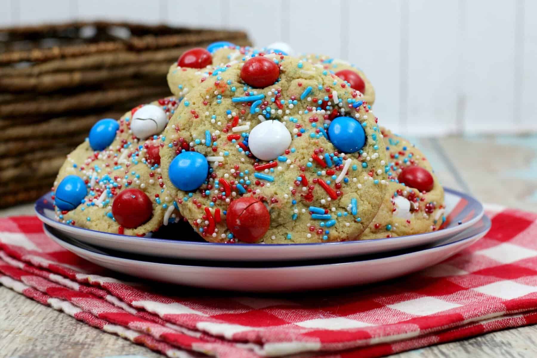 A plate of cookies decorated with red, white, and blue candies and sprinkles sits on a red and white checkered cloth, with a rustic basket and wooden surface in the background.