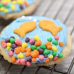 A round sugar cookie with blue frosting, topped with two fish-shaped crackers and colorful candy pieces, creates an underwater scene—perfect as a fun summer snack for kids. The cookie rests on a metal cooling rack.