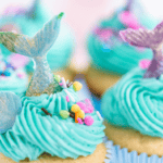 Cupcakes topped with turquoise frosting, colorful sprinkles, and decorative mermaid tails and seashells are perfect summer snacks for kids, creating a fun ocean-themed appearance.