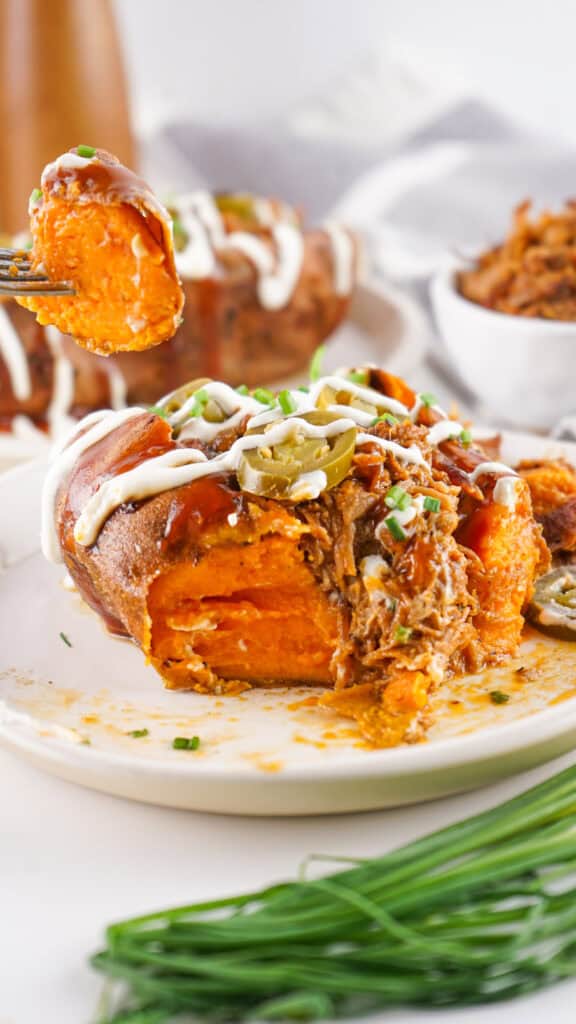 A baked sweet potato topped with shredded meat, sliced jalapeños, chopped chives, and drizzled with a white sauce sits on a plate. A fork holds a bite-sized piece above the plate.