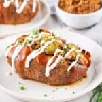 A baked sweet potato topped with shredded meat, sliced jalapeños, chives, and drizzled with white sauce sits on a white plate—a delicious idea for summer sweet potato recipes. A bowl of rice and another plated potato are in the background.