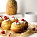 Mini pineapple upside-down cakes, perfect as mini desserts, are topped with whipped cream and maraschino cherries on brown parchment paper, with a whole pineapple and stacked white plates in the blurred background.