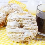 Three crumb-topped donuts drizzled with white icing sit on a yellow checkered cloth next to a glass cup of black coffee, making the perfect treat for a cozy Mother's Day brunch. A small glass pitcher is in the background.