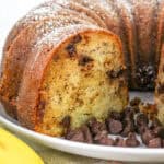 A sliced bundt cake with chocolate chips inside, dusted with powdered sugar, sits on a white plate. Perfect for those seeking delicious ripe banana recipes, chocolate chips and a banana are placed nearby.
