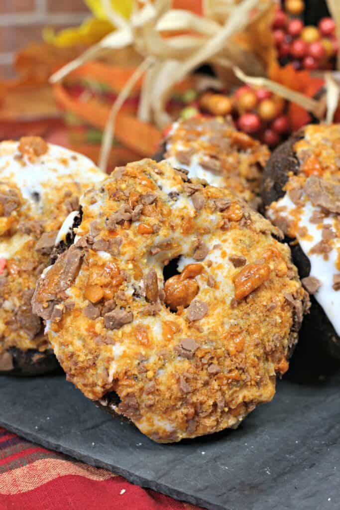 Three homemade donuts covered with white icing and crushed toffee pieces are displayed on a dark tray, with autumn-themed decorations and berries blurred in the background.