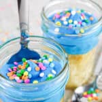 Two glass jars filled with yellow cake and topped with bright blue frosting and colorful round sprinkles—perfect birthday cakes ideas. One jar has a spoon in it; the other sits behind, surrounded by scattered sprinkles.