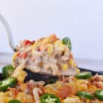 A metal spatula lifts a serving of cheesy cowboy pasta recipe casserole with ground meat, corn, diced tomatoes, and sliced jalapeños from a skillet. The casserole is topped with melted cheese and extra jalapeño slices.