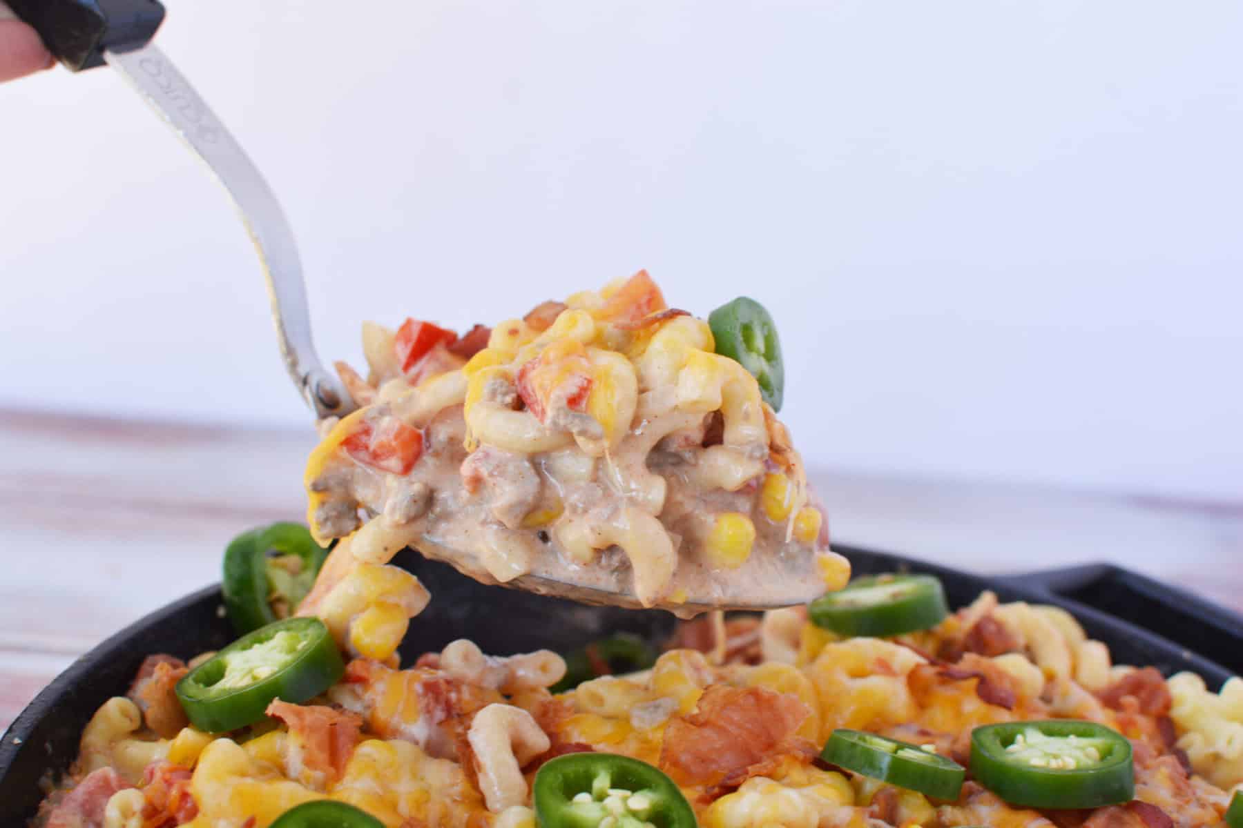A metal spatula lifts a serving of cheesy cowboy pasta recipe casserole with ground meat, corn, diced tomatoes, and sliced jalapeños from a skillet. The casserole is topped with melted cheese and extra jalapeño slices.