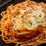 A close-up of a classic chicken parmesan recipe topped with melted cheese and herbs, served on a bed of spaghetti with tomato sauce on a dark plate.