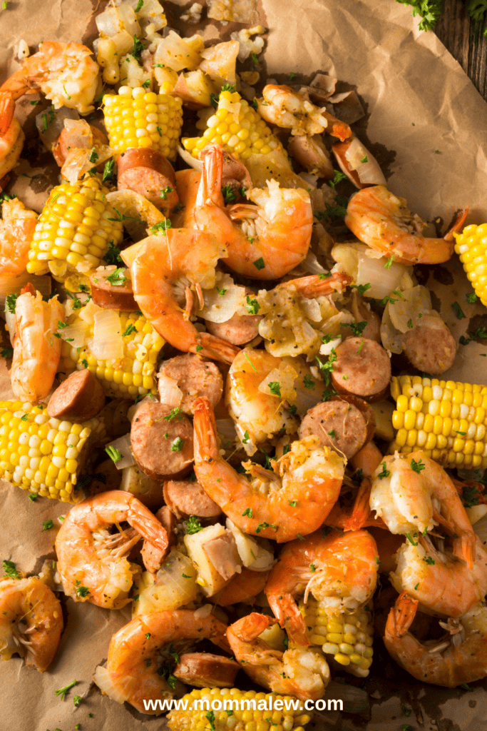 A close-up of a Seafood Boil featuring shrimp, sausage slices, corn on the cob, and potatoes, all seasoned and garnished with herbs, spread out on brown parchment paper.