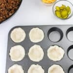 A muffin tin with dough pressed into each cup, ready to make cheeseburger biscuit cups, sits beside a skillet of cooked ground beef, a bowl of shredded cheddar cheese, and a bowl of sliced pickles on a white surface.