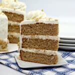 A slice of three-layer cinnamon cake with white frosting sits on a small plate in front of the whole cake. Both are decorated with frosting and crumbs, making them perfect winter cakes, with plates stacked in the background on a blue plaid cloth.