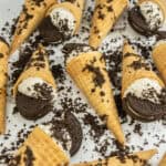 Oreo Cheesecake Cones filled with cookies and cream dessert and topped with crushed chocolate sandwich cookies are arranged on a white surface, with cookie crumbs scattered around.
