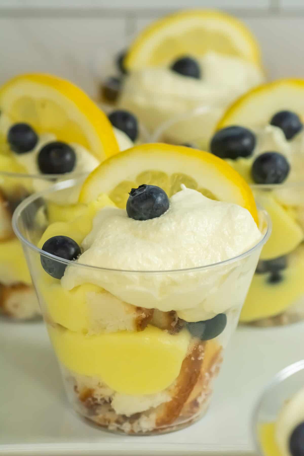 A clear cup filled with layers of lemon pudding, cake pieces, whipped cream, blueberries, and topped with a lemon slice and more blueberries—these mini lemon blueberry trifles are joined by other delicious dessert cups in the background.
