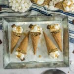 Five waffle cones filled with mini marshmallows, chocolate pieces, and crushed graham crackers are arranged on a metal tray—an irresistible twist for anyone seeking Unique S’mores Ideas. Nearby, a bowl of marshmallows and chocolate squares rests on a striped cloth.