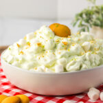 A white bowl filled with creamy key lime pie fluff dessert topped with mini marshmallows and a vanilla wafer, sitting on a red checkered cloth with more wafers and marshmallows around it; a plant is in the background.