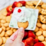 A hand holds a vanilla wafer topped with creamy pistachio dip. In the background, a platter displays more wafers, cookies, strawberries, and a bowl of dip garnished with whipped cream and a cherry.