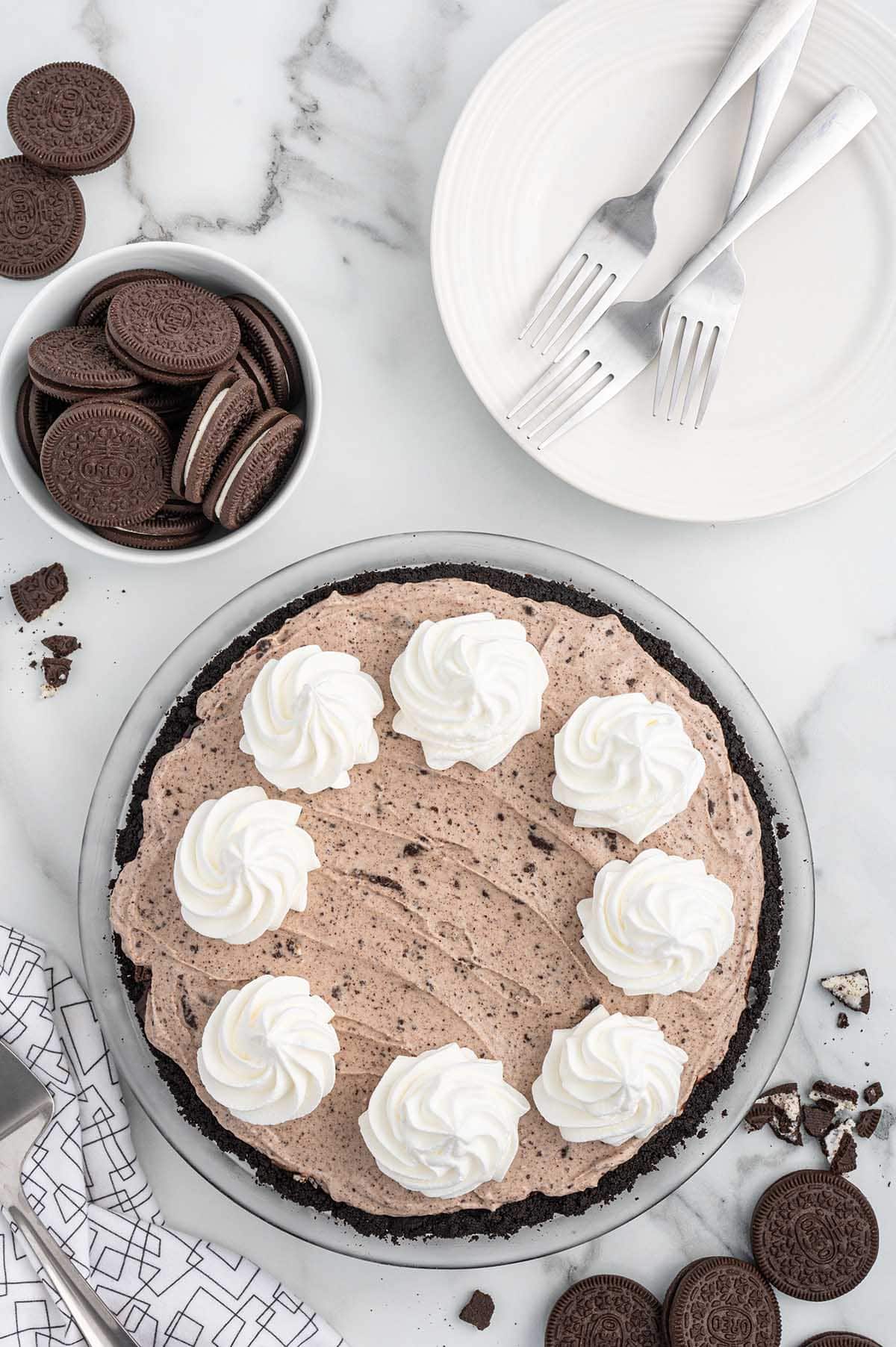 A chocolate cream pie with an Oreo cookie crust, topped with dollops of whipped cream. Nearby are a bowl of Oreo cookies, a plate with two forks, and a napkin, all on a white marble surface.