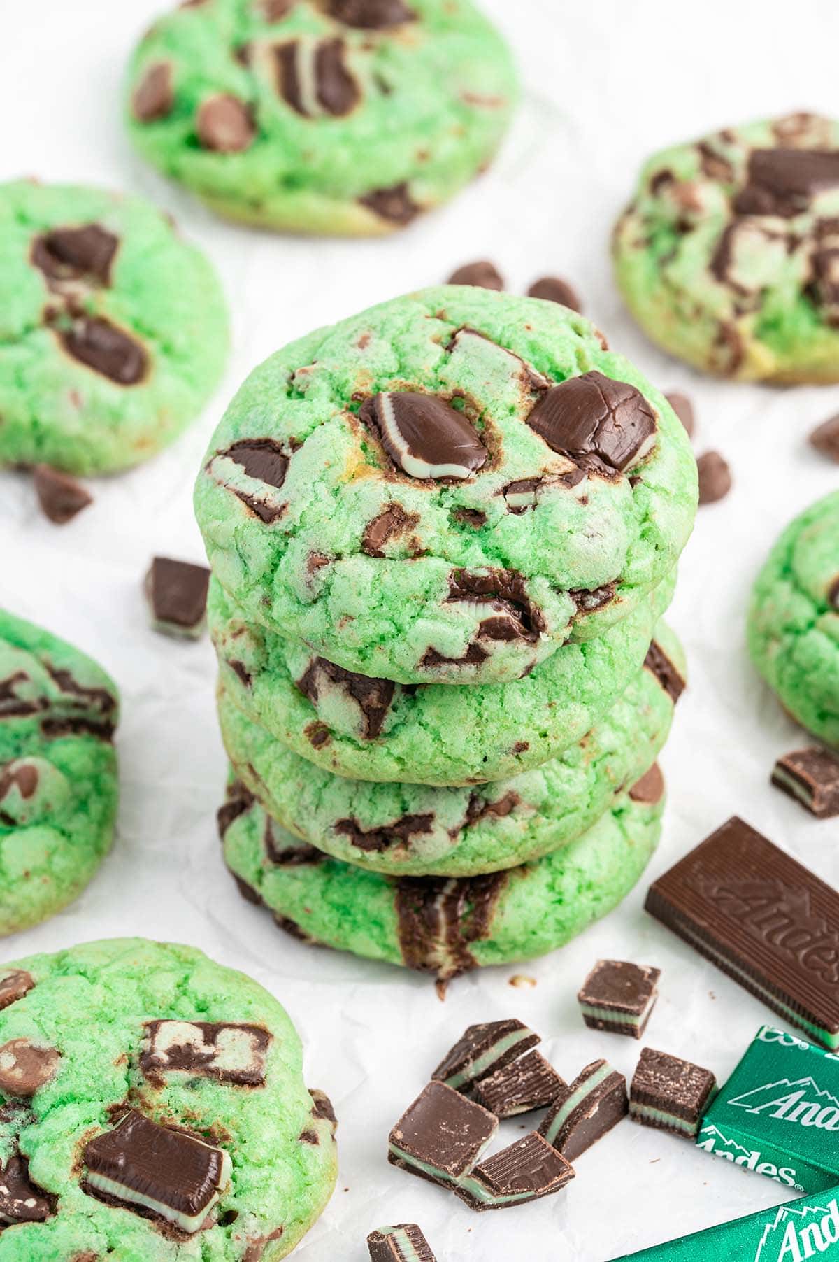 A stack of green cookies with chunks of chocolate and mint, inspired by delicious chocolate chip cookie recipes, surrounded by more cookies and pieces of mint chocolate on a white surface.