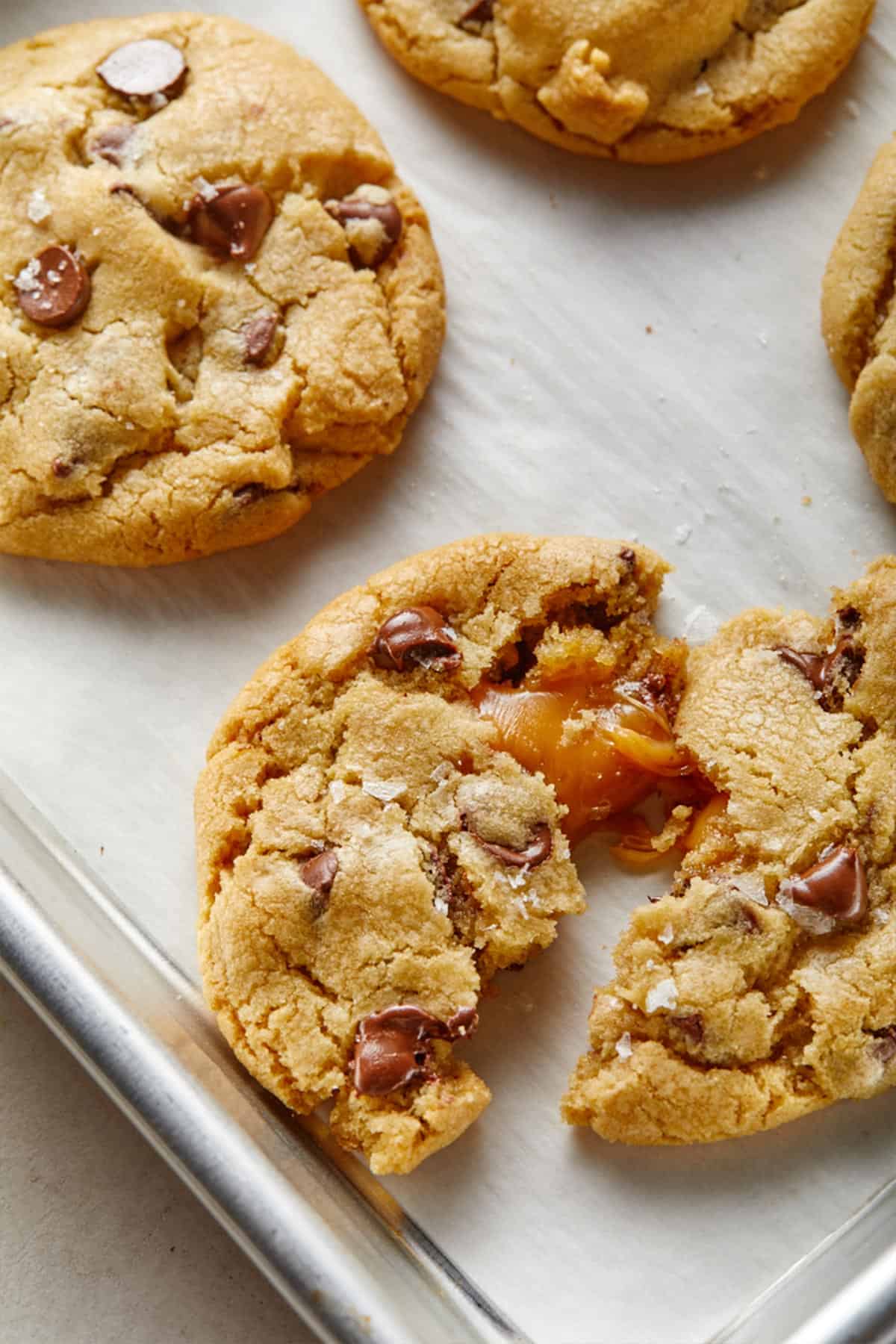 A close-up of chocolate chip cookies on a baking sheet, with one cookie broken in half to reveal gooey caramel inside—perfect inspiration for new chocolate chip cookie recipes.