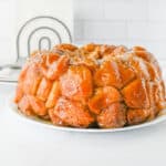 A glazed monkey bread, a creative twist on desserts with biscuits, sits on a white plate, showcasing its golden-brown, pull-apart pieces. The background features stacked plates, forks, and a white towel with a black circular design.