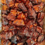 Chunks of browned, caramelized meat, possibly pork burnt ends, arranged closely together in a foil pan and coated with a rich, shiny glaze—a delicious result of learning how to make burnt ends in the oven.