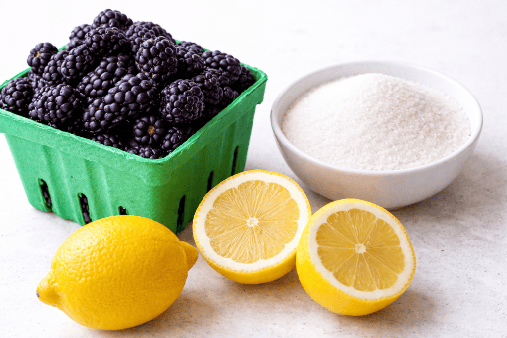 A green carton of fresh blackberries, a white bowl filled with granulated sugar, one whole lemon, and one lemon cut in half arranged on a white surface—everything you need to make homemade blackberry simple syrup.