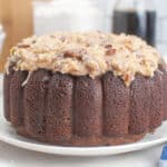 A round chocolate bundt cake topped with a thick layer of coconut pecan frosting sits on a white plate—perfect for those seeking Easter dessert ideas, with bowls and kitchen items blurred in the background.