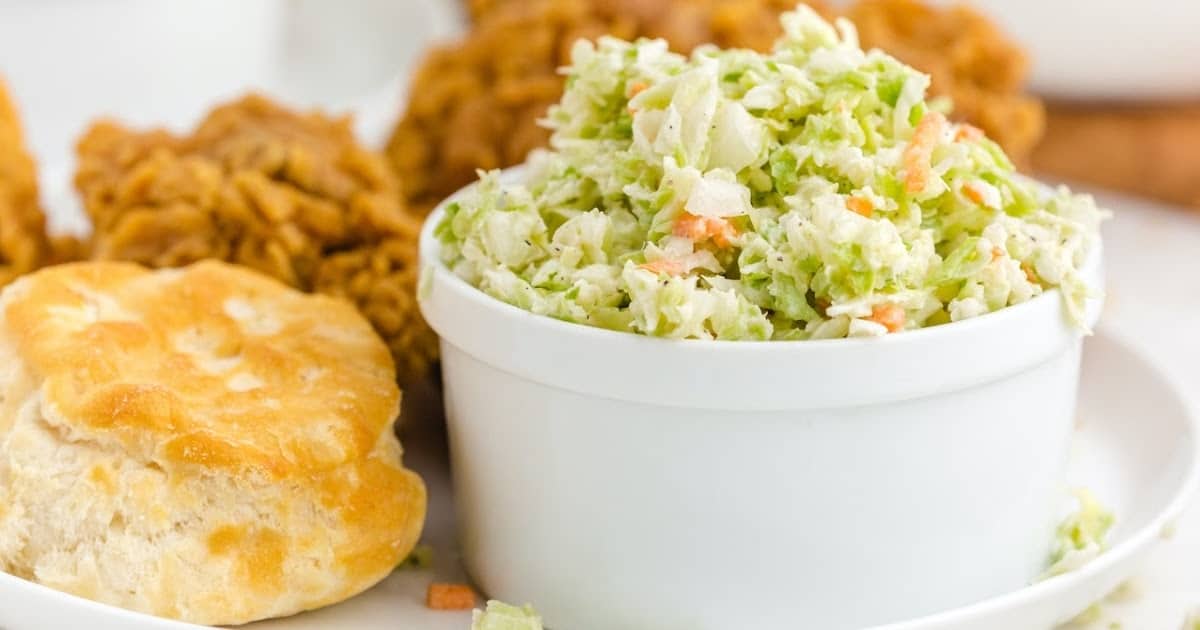 A white bowl filled with coleslaw sits on a plate next to a flaky biscuit and pieces of fried chicken—perfect companions for classic burnt ends side dishes.