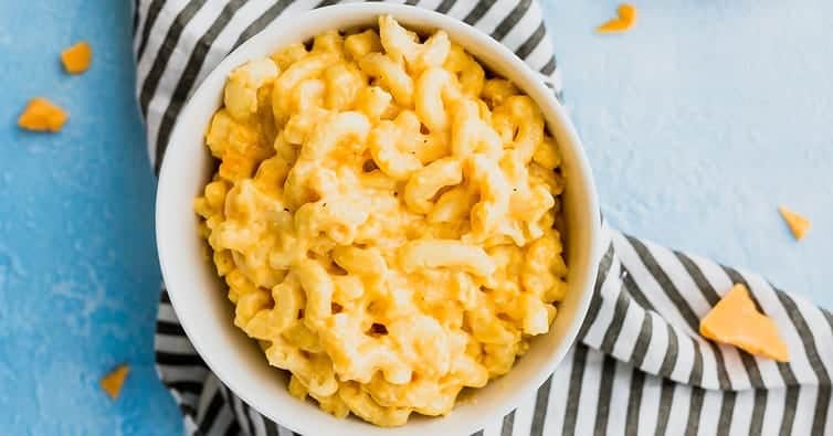 A white bowl filled with creamy macaroni and cheese sits on a black and white striped cloth, with a light blue background and small pieces of cheddar cheese nearby.