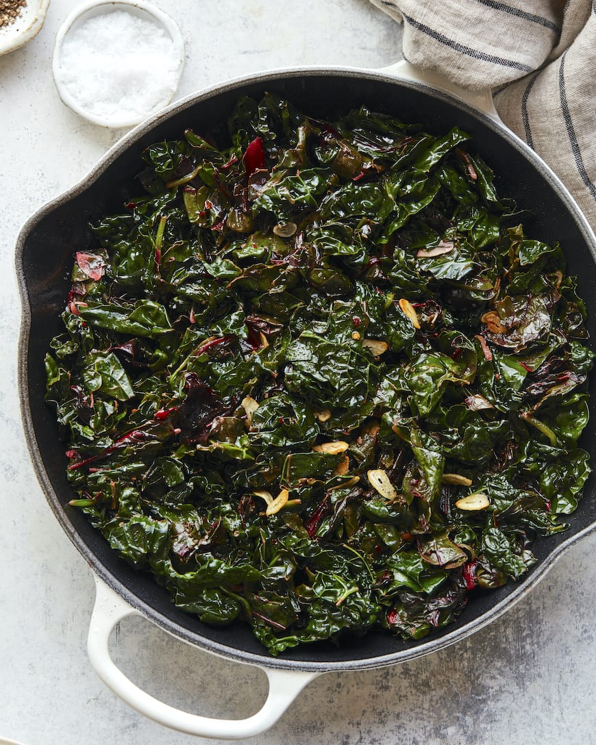 A skillet filled with sautéed Swiss chard and sliced garlic, glistening from cooking. Perfect as one of the side dishes for a shrimp boil, it sits beside a dish of coarse salt and a striped towel on a light surface.
