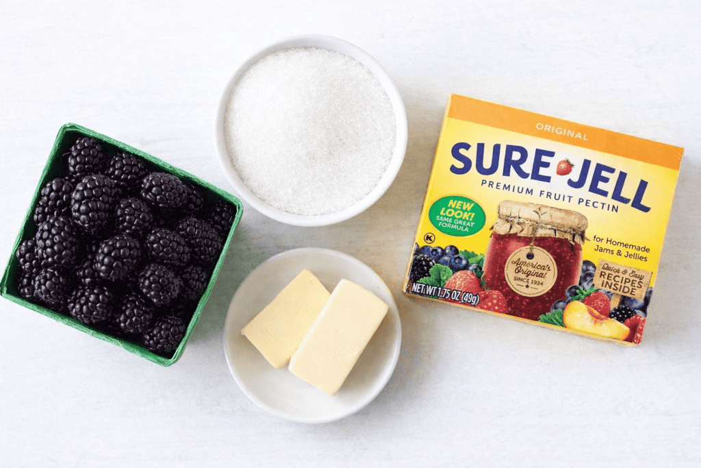 A carton of fresh blackberries, a bowl of granulated sugar, two slices of butter, and a box of Sure-Jell premium fruit pectin arranged on a white surface—everything you need for a delicious blackberry jam recipe.