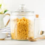 A large glass jar filled with brown sugar sits on a white countertop, ready for anyone curious about how to make brown sugar. Nearby are a small bowl, wooden spoon, striped cloth, white canister, measuring cups, and a jug with white flowers.