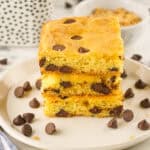 A plate with a stack of three chocolate chip cookie bars—one of many delicious uses for yellow cake mix—is surrounded by scattered chocolate chips, with a fork, spoon, and a small bowl of brown sugar in the background.