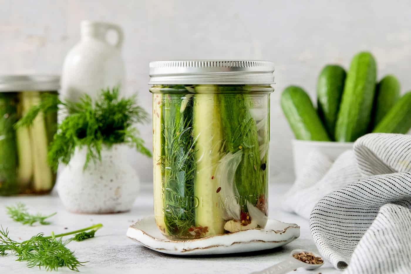 A jar of homemade pickles with fresh dill and garlic sits on a small white plate—perfect as one of your burnt ends side dishes. In the background are cucumbers in a bowl, a vase with dill, and a striped cloth, all on a light surface.
