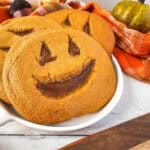 Three large pumpkin-shaped cookies with smiling jack-o-lantern faces sit in a white dish. Perfect for a Halloween Dessert Table, theyre surrounded by a plaid orange cloth and decorative gourds for a festive autumn vibe.