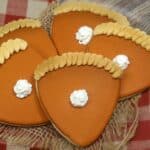 Five pumpkin pie sugar cookies decorated with crust edges and a dollop of white icing in the center, arranged on a rustic wooden surface with burlap and a red plaid cloth.