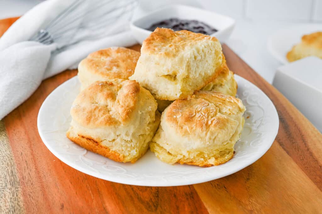 A white plate filled with golden, flaky biscuits sits on a wooden board—perfect for christmas brunch ideas. In the background, a dish of dark jam and a white napkin with a whisk can be seen.